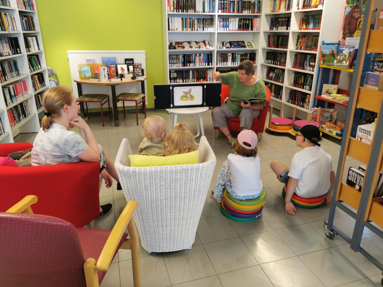 Nina beim Vorlesen: Nina sieht man von vorne und die am Boden und auf einem Sessel sitzenden Kinder von hinten. An den Seiten Bücherregale. Und auch hinter Nina Bücherregale.