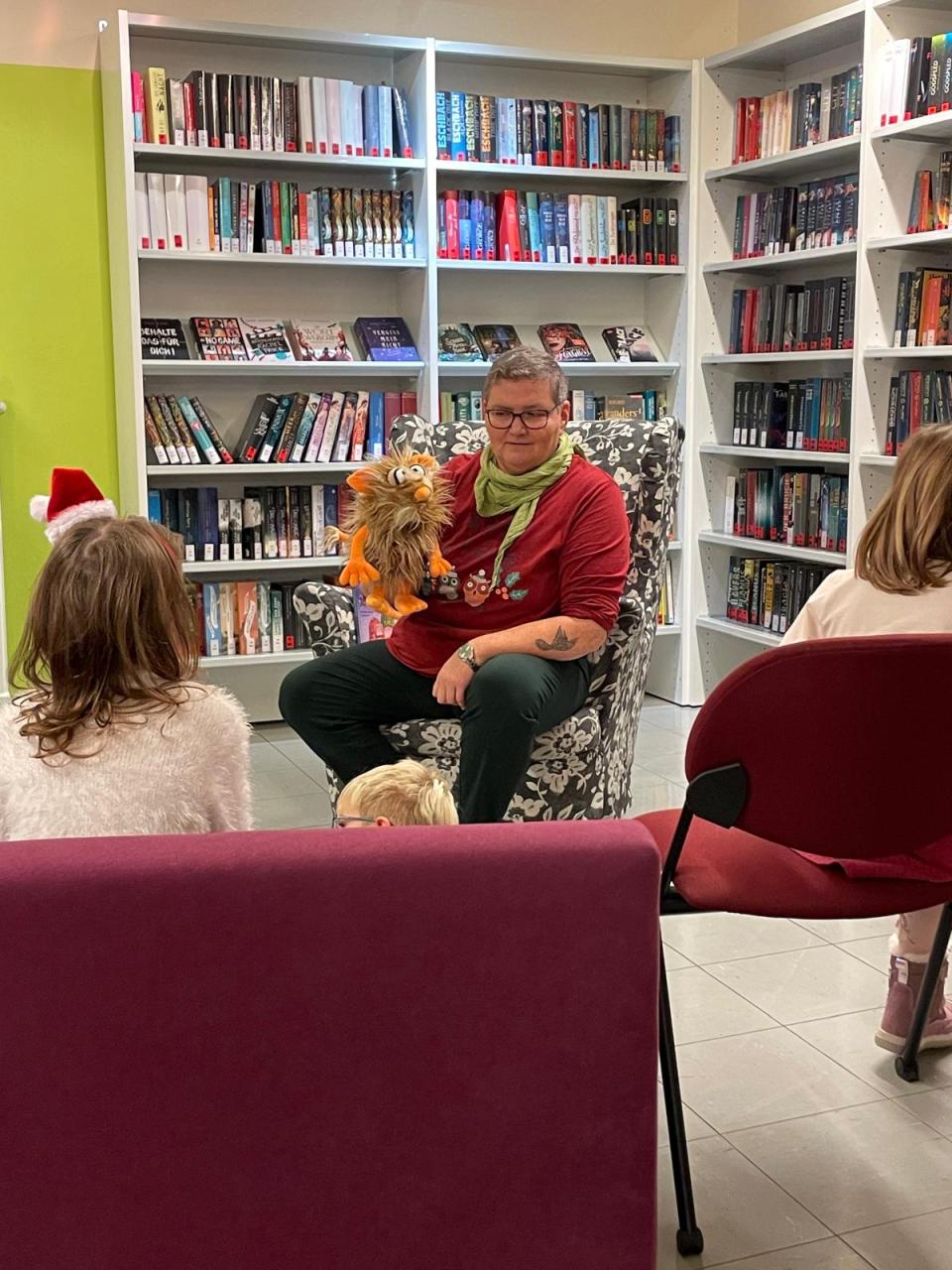 Mitarbeiterin Nina beim Vorlesen. Man sieht sie von vorne und ein paar Kinder auf Stühlen von hinten. Nina sitzt vor Bücherregalen
